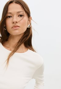 Top bianco a maniche lunghe con scollatura rotonda, texture liscia e design aderente. La modella indossa orecchini rotondi, audaci e riflettenti.