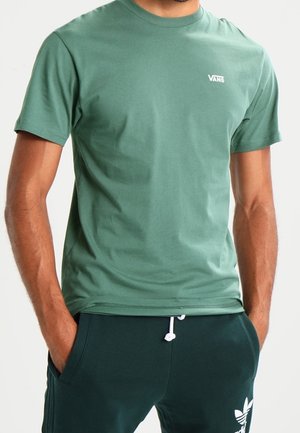 Hombre con camiseta verde de Vans y pantalones deportivos Adidas verde oscuro con las manos en los bolsillos, de pie contra un fondo liso.