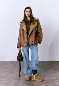 Chaqueta marrón de piel vuelta con gran cuello, combinada con camiseta de rayas, jeans anchos de mezclilla y botas color cuero con detalles de costuras en contraste.