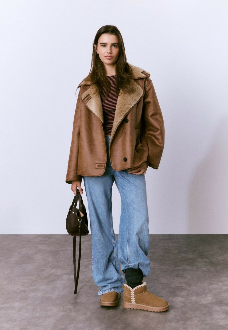 Chaqueta marrón de piel vuelta con gran cuello, combinada con camiseta de rayas, jeans anchos de mezclilla y botas color cuero con detalles de costuras en contraste.