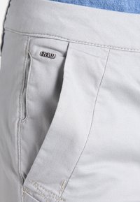 Pantaloni corti grigio chiaro con un tessuto liscio e testurizzato, tasca laterale e un'etichetta metallica "RAW" in accentuazione sulla vita.