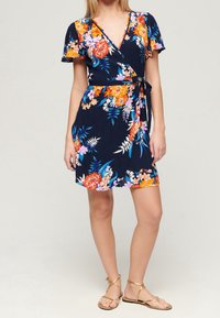 Blommig omslagsklänning med marin bakgrund, prydd med livfulla orange, rosa och vita blommor. Korta ärmar och ett knyt i midjan.