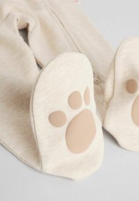 Un body color beige claro con cierre de cremallera y detalles con forma de patas en los pies, con almohadillas beige. Tejido suave con una apariencia texturada.