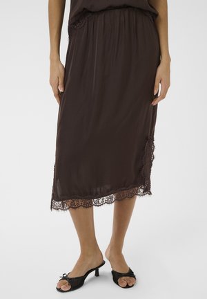 Femme portant une jupe midi marron foncé avec un ourlet en dentelle, associée à des mules noires à talons bas ouverts à l'avant avec des nœuds, sur un fond uni.