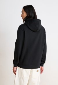 Felpa nera con cappuccio, dotata di polsini e fondo a costine, abbinata a pantaloni beige con un'etichetta del marchio ben visibile sulla tasca posteriore.