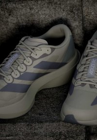 Ζευγάρι ανοιχτό μπεζ παπούτσια Adidas Adizero για τρέξιμο με σκουρόγριμες ρίγες και χονδρές σόλες σε επιφάνεια με υφή πέτρας.