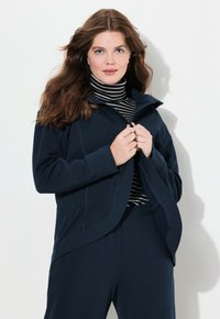 Mörkblå zip-up fleecejacka med hög krage, tillsammans med en svart och vit randig turtleneck. Mjuk textur, avslappnad passform och minimala sömmar.