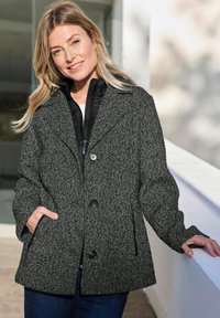 Lachende vrouw met lang blond haar draagt een gestructureerde grijze jas met knopen en een donkere binnenjas, staand buiten bij een witte leuning.