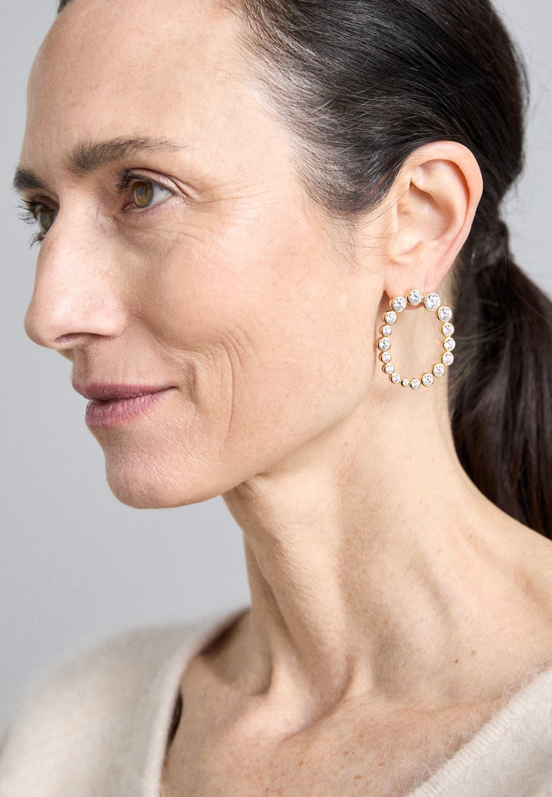 Χρυσά σκουλαρίκια κρίκοι σε σχήμα ανοιχτού οβάλ, στολισμένα με πολλούς μικρούς διαφανείς ρουμπίνους, που αντανακλούν το φως σε ουδέτερο φόντο.