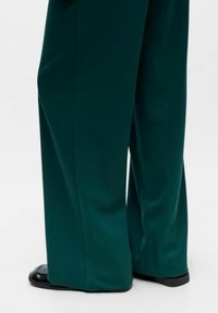 Wide-leg trousers made of smooth, dark teal fabric. Features a high waist and straight silhouette with minimal seams; paired with black shoes.