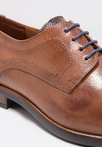 Braune Lederschuhe mit glatter Textur, detaillierter Nähte und einem gemusterten dunkelbraunen Akzent. Mit blauen Schnürsenkeln und einem Holzabsatz.