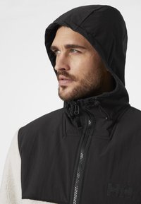 Chaqueta negra con capucha y cuello alto, que cuenta con un cierre de cremallera en el frente, cordones elásticos y un suave acento de forro polar blanco en un lado.