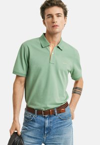 Polo verde con colletto arancione, maniche corte e un logo sul petto. Indossato con jeans blu e una cintura intrecciata marrone.