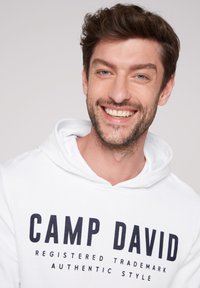 Witte hoodie met een groot zwart gedrukt logo "CAMP DAVID" en kleinere tekst eronder. Zachte stof en trekkoord bij de hals, casual pasvorm.