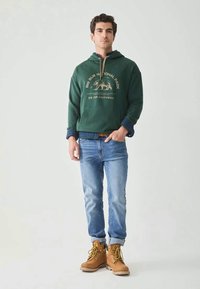 Jonge man in een groene Big Sur-hoodie, opgerolde lichtblauwe jeans, bruine laarzen, staand met één hand in zijn zak tegen een effen achtergrond.