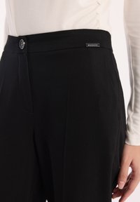 Zbliżenie na osobę ubraną w czarne spodnie o wysokim stanie z guzikiem i metką marki Makalu, w połączeniu z jasnym długim rękawem.