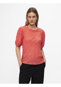 Koraalkleurige blouse met pofmouwen, voorzien van ingewikkelde kant oogjes en een ronde halslijn. Gecombineerd met zwarte broek voor contrast.