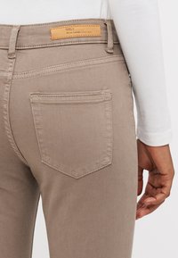 Beige denim jeans med nærbilde av en baklomme og lærmerke på midjebåndet. Har en jevn tekstur og en tettsittende design.