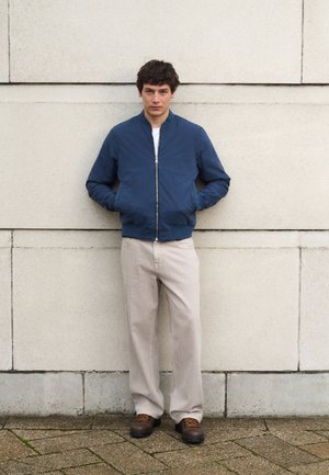 Nuori mies seisoo beigeä seinää vasten, pukeutuneena siniseen vetoketjutakkiin, valkoiseen paitaan, vaaleanbeigeihin housuihin ja ruskeisiin nauhallisiin kenkiin.