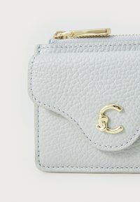 Porte-cartes en cuir texturé gris clair avec un accent en métal doré portant un logo. Fermeture éclair en haut et bords cousus.