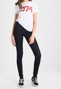 Vit t-shirt med röd "1974" grafik, kombinerad med mörka skinny jeans och svarta sneakers med vita sulor, vilket skapar en avslappnad outfit.