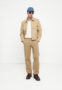 Cream-colored jacket with two front pockets, paired with a white T-shirt and beige trousers. Blue denim cap and black sunglasses complete the look.