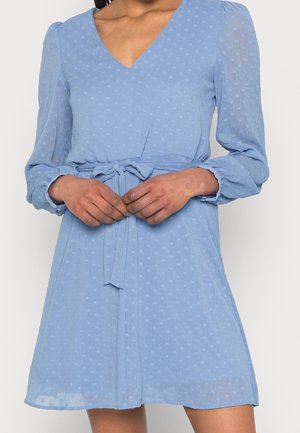 Femme portant une robe bleu clair à manches longues texturée, avec un col en V et une ceinture nouée à la taille, les mains délicatement jointes devant.