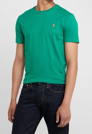 Man draagt een groen T-shirt met korte mouwen met een klein oranje geborduurd logo op de linkerborst en donkerblauwe spijkerbroek.