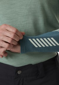 Rukávník s dlouhými rukávy v temně modré tkanině s texturovaným vzorem, zdobený bílými diagonálními pruhy na manžetě. Nosený přes světle zelený top.