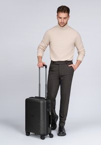 Hombre con un suéter de cuello alto beige y pantalones oscuros caminando con una maleta negra con ruedas, una mano en el bolsillo, fondo neutro.