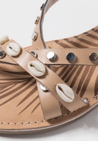Sandal de color bronce con correas de cuero, tachuelas plateadas y adornos de conchas de cowrie blancas sobre una plantilla con un patrón.
