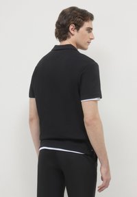 Giovane uomo di profilo che indossa una polo nera a maniche corte sopra una camicia bianca e pantaloni neri su uno sfondo neutro.