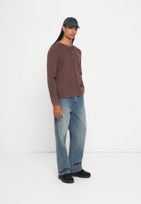 Person, der ein braunes Langarm-Henley-Shirt, weit geschnittene, ausgebleichte blaue Jeans, schwarze Sneaker und eine schwarze Mütze trägt, und mit einer Hand in der Tasche steht.