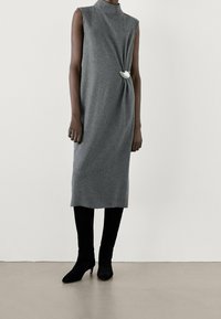 Graues ärmelloses Kleid mit hohem Kragen, mit einer verdrehten Detail und einem weißen Akzent. Kombiniert mit knielangen schwarzen Stiefeln.