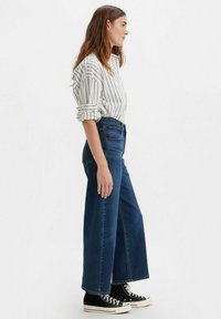 Vaqueros de pierna ancha en azul oscuro, combinados con una blusa blanca a rayas. La modelo lleva zapatillas altas negras. Diseño sencillo, ajuste casual.