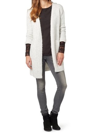 Helles grauer langer Cardigan aus einem weichen, strukturierten Stoff, mit Seitentaschen und offener Vorderseite. Getragen über einem dunklen Oberteil und grauen Skinny Jeans.