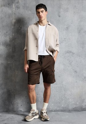 Fiatal férfi szürke fal előtt áll, bézs gombos inget visel fehér póló felett, barna cargo rövidnadrággal, fehér zoknival és bézs tornacipővel.