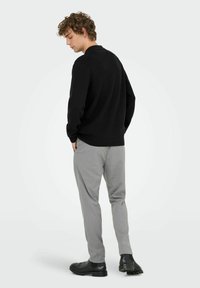 Schwarzer Strickpullover, graue maßgeschneiderte Hose und schwarze Stiefel. Das Modell steht mit dem Rücken zur Kamera und zeigt eine lockere Passform sowie glatte Texturen.
