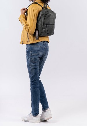 Czarny plecak skórzany o teksturowanej powierzchni, z metalowymi elementami w kolorze srebrnym i regulowanymi ramiączkami. Noszony na żółtej kurtce i niebieskich dżinsach.