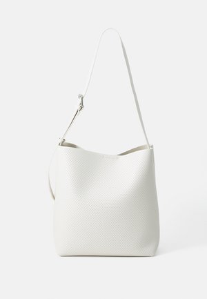 Sac en cuir perforé blanc avec une bandoulière ajustable et une boucle en argent, présenté sur un fond blanc uni.