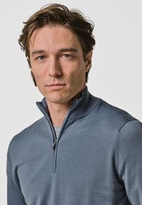 Sweater zippé bleu clair avec un col haut, fabriqué en tissu tricoté texturé, doté d'un léger brillant et d'une coupe ajustée.