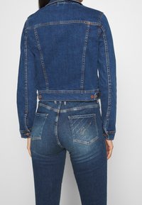 Džínová bunda a džíny tmavě modré barvy. Bunda má klasický střih s špičatým límcem, knoflíkovým zapínáním a zadní vsadkou. Džíny mají dekorativní prošití.