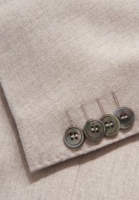 Ljust brunt tyg med en slät textur, med fyra mörka, runda knappar med fyra hål och subtila sömnadsdetaljer ovanför knapparna.