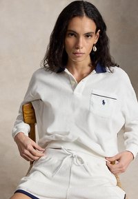 Sötét, göndör hajú nő ül egy fából készült széken, fehér hosszú ujjú galléros ingben és fehér rövidnadrágban, tengerészkék szegéllyel.