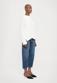 Pull en tricot blanc avec un col rond et des fentes sur les côtés, associé à un jean en denim bleu ample et des escarpins noirs à bout pointu.