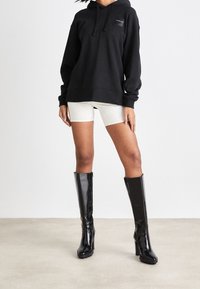 Svart oversized huvtröja med logga fram, i kombination med beige cykelbyxor och knähöga blanka svarta klackstövlar.