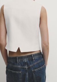 Biały top bez rękawów o skróconym kroju z rozcięciem z tyłu, zestawiony z ciemnoniebieskimi szortami dżinsowymi z elastycznym paskiem w talii i tylniami kieszeniami.