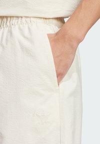 Light beige athletic pants with a textured fabric, elastic waistband, and side pockets; features an embroidered Adidas logo near the hip.