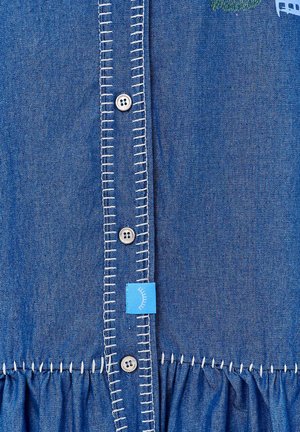 Džínová košile s modrou tkaninou, bílým kontrastním šitím podél lišty, stříbrnými knoflíky a nabranou látkou na lemu.