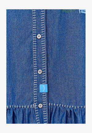 Koszula dżinsowa z niebieskiego materiału, białym kontrastowym szwem wzdłuż zapinania, srebrnymi guzikami oraz marszczonym materiałem u dołu.